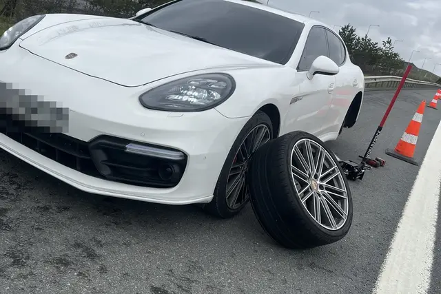 Yol kenarında bir binek aracın sökülmüş tekerleği yere yatırılmış; kriko aracın altında, arkada güvenlik konileri sıralı.